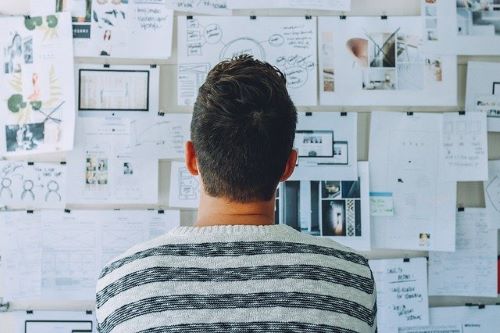 The back of a man stood in front a wall containing pieces of paper with different diagrams and pictures. 