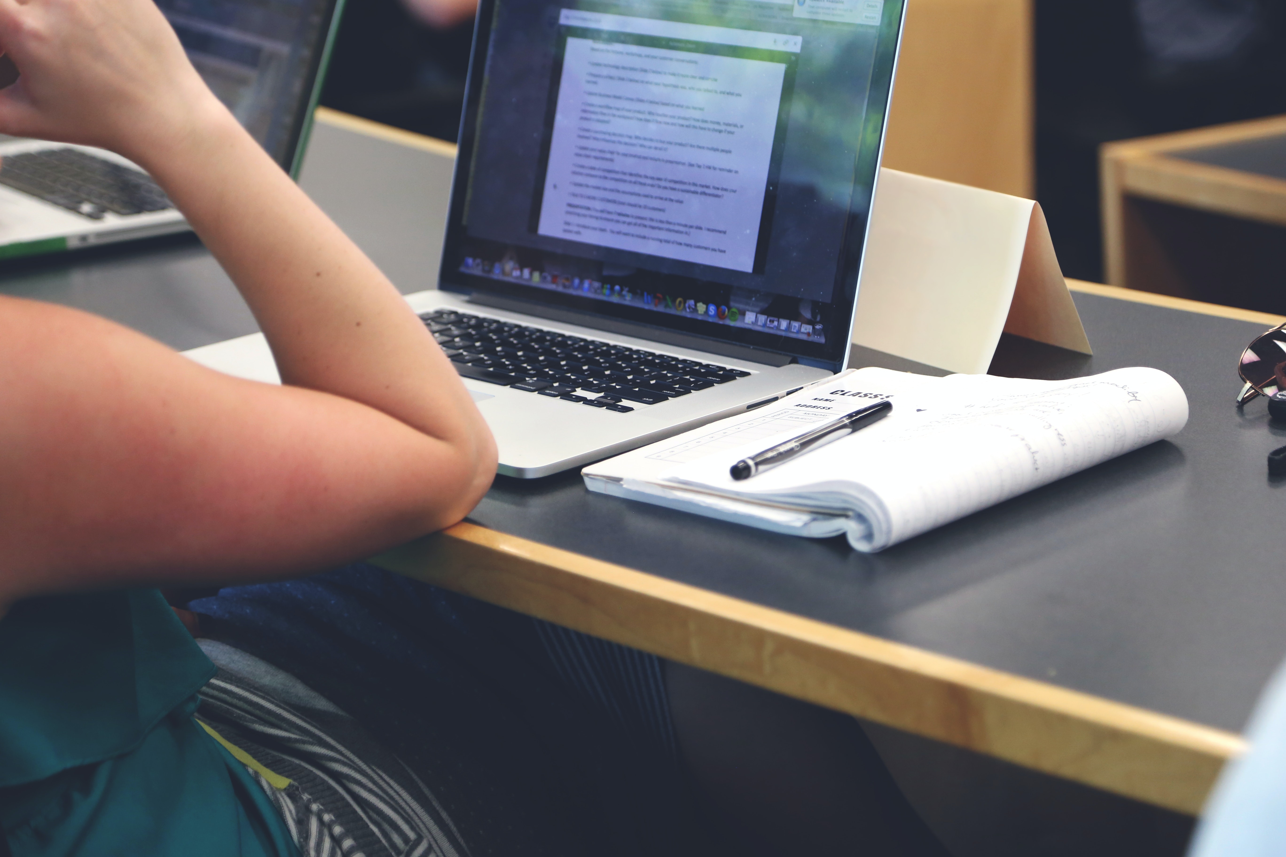 Person sat at a laptop, with a notebook and pen by the side of it. 